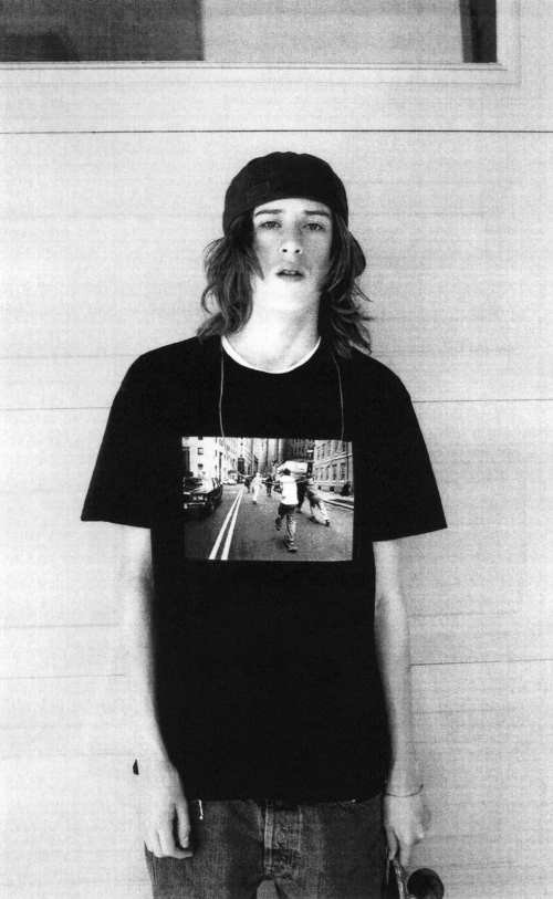 Young adult wearing a black graphic tee shirt and a beanie, standing in front of a gray paneled wall, holding a skateboard.