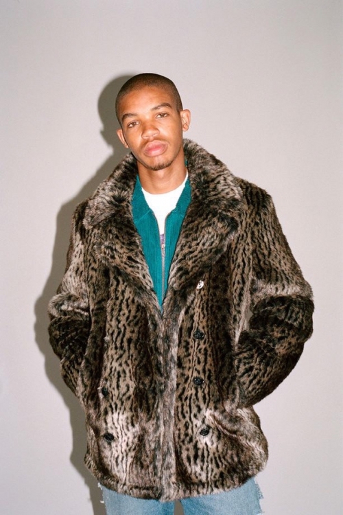 Man in faux fur coat, wearing a teal jacket and jeans, posing in a photo studio with even lighting against a pale gray background.