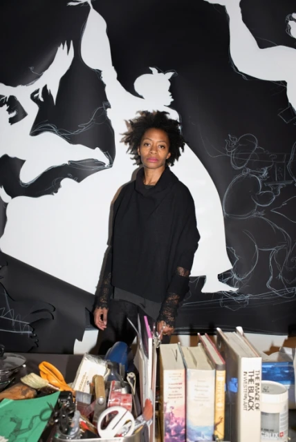 Adult woman posing in front of a black and white mural with artwork and books in foreground, artistic environment, dark clothes.