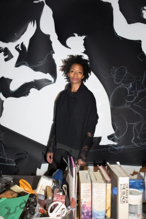 Adult woman posing in front of a black and white mural with artwork and books in foreground, artistic environment, dark clothes.