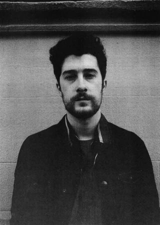 Black and white portrait of a man with a beard and dark hair wearing a dark jacket against a plain textured wall in New York.