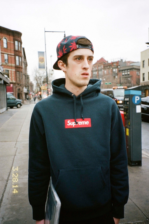 A young man in a Supreme hoodie and hat stands on a city sidewalk with buildings and cars in the background, captured in a medium shot.