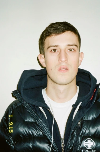 Young man in a puffy jacket with a hood poses indoors against a plain background, looking directly at the camera in a medium shot.