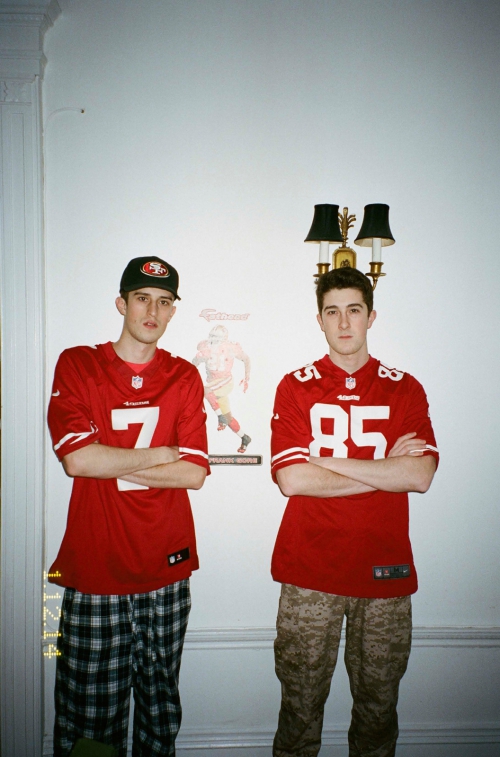 Two adult males in jerseys stand indoors, posing with arms crossed in front of white walls, indoor lighting, and a portrait hanging nearby.