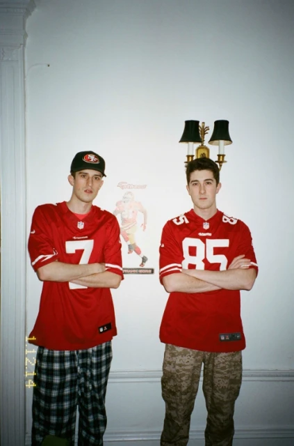 Two adult males in jerseys stand indoors, posing with arms crossed in front of white walls, indoor lighting, and a portrait hanging nearby.