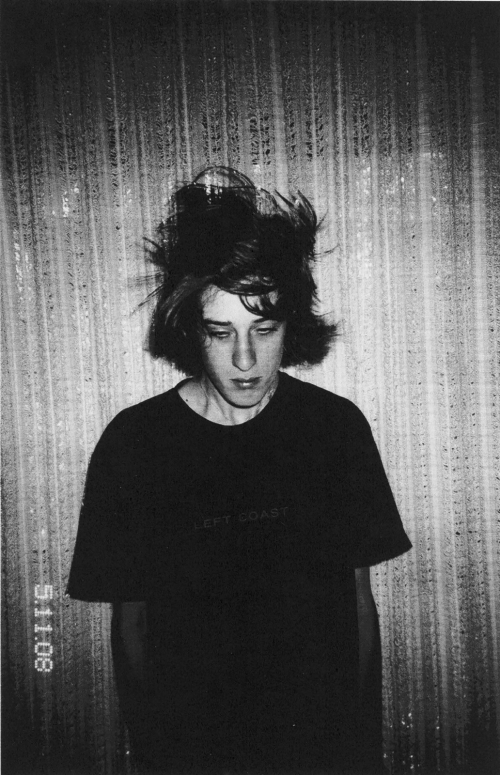 A young adult with tousled hair stands in front of a textured backdrop, wearing a dark T-shirt in a black and white portrait.