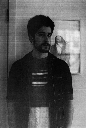 Portrait of a man with dark hair and a beard in front of a framed art print. He wears a striped shirt and open button-down.