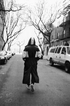 Monochrome street-level photograph of a figure walking away down a New York City street lined with parked cars and bare trees.
