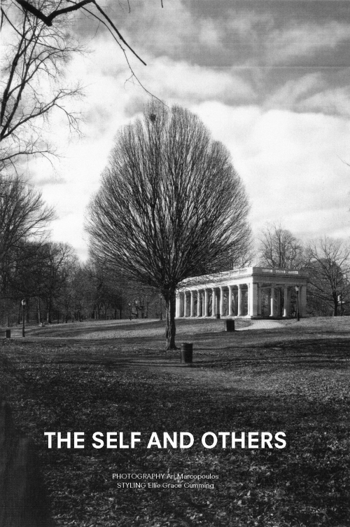 Monochrome outdoor photograph of a tall tree centered in a park-like setting with a colonnade structure in the background under a cloudy sky.
