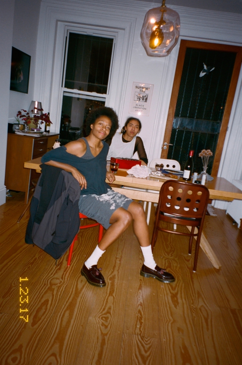 Two people seated at a wooden dining table inside a residential room with hardwood floors, photographed by Ari Marcopoulos.