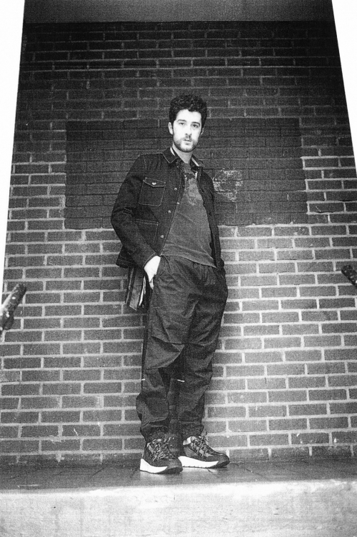 Black and white portrait of a young adult man in jacket and casual pants leaning against a brick wall on a step outside.