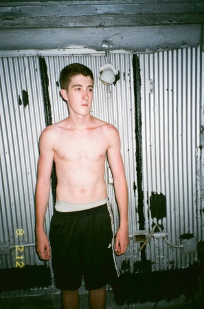 Shirtless young man standing against a corrugated metal wall wearing black athletic shorts with white side stripes.