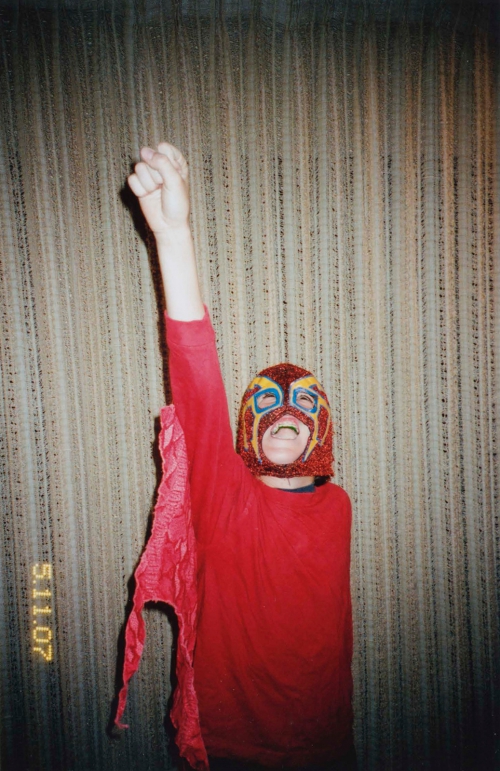 Person wearing a red shirt and a colorful wrestling mask raising an arm in a room with light brown curtains in the background.