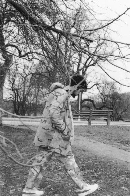 Adult male in camouflage apparel walks in a park with bare trees and a wooden bench in the background, captured in a monochrome photo.