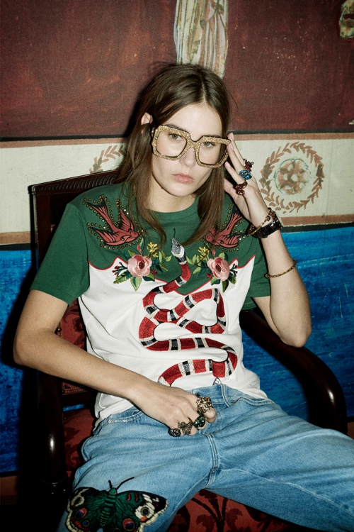 Woman with glasses wearing a t-shirt with a snake and bird design, and jeans with a butterfly patch sitting indoors.