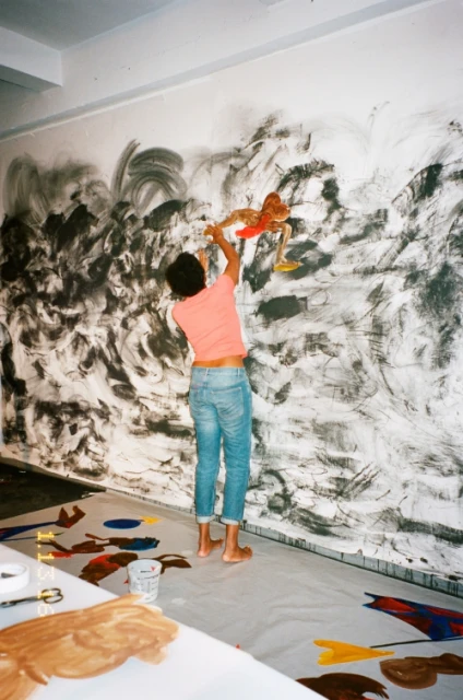 A woman is seen from the back, applying black paint to a white wall in a brightly lit indoor space with splattered paint on the floor.