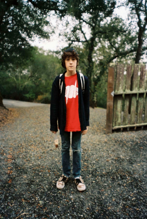 Teenager stands on a gravel path wearing jeans, a graphic tee, and a black jacket against a background of trees and an old wooden gate.