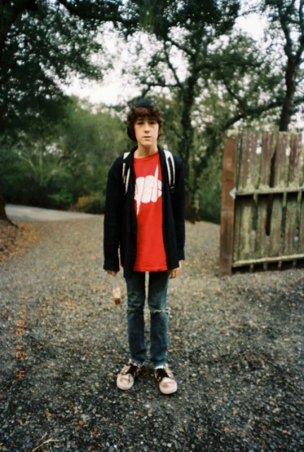 Teenager stands on a gravel path wearing jeans, a graphic tee, and a black jacket against a background of trees and an old wooden gate.