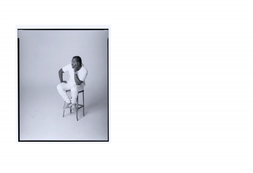 Monochrome portrait of adult Black male model posing while sitting on stool in a photo studio with soft lighting and relaxed facial expression.