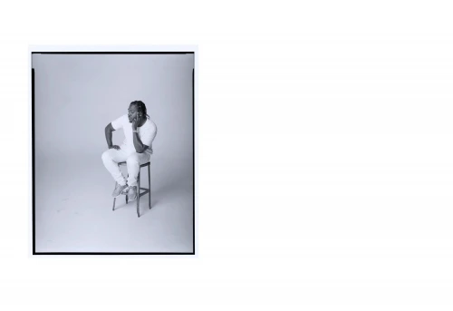 Monochrome portrait of adult Black male model posing while sitting on stool in a photo studio with soft lighting and relaxed facial expression.