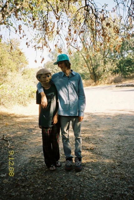 Two people pose in an outdoor setting; one is wearing an E.T. mask, while the other wears a teal cap and has an arm around the mask wearer.
