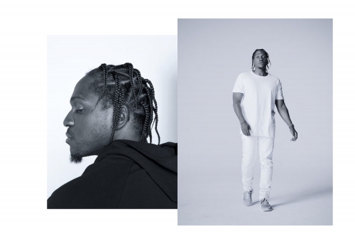Black and white studio portrait of a male with braids, wearing a white shirt and jeans, standing against a clean backdrop.