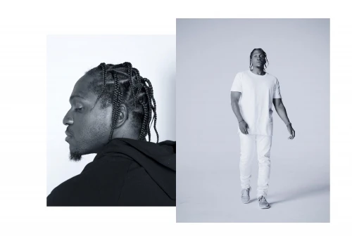 Black and white studio portrait of a male with braids, wearing a white shirt and jeans, standing against a clean backdrop.