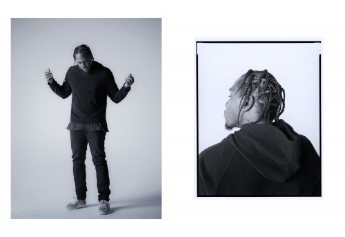 Black and white portrait of an adult male with braids, wearing a dark sweatshirt and jeans in a photo studio with soft lighting.