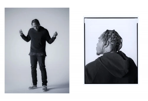 Black and white portrait of an adult male with braids, wearing a dark sweatshirt and jeans in a photo studio with soft lighting.