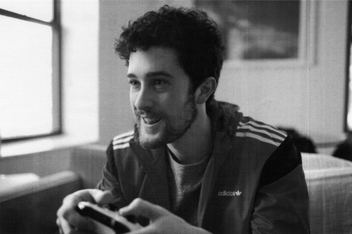 Young adult male smiling and holding a gaming controller indoors, dressed in a vintage-style Adidas jacket near a window in black and white.