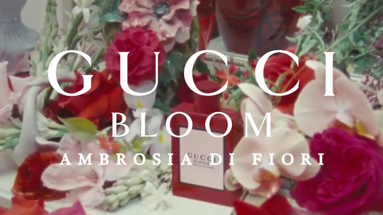 Close-up of Gucci Bloom Ambrosia di Fiori perfume bottle surrounded by an arrangement of colorful roses and orchids.