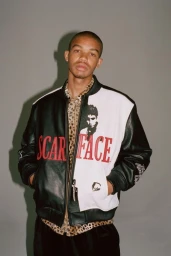 Portrait of a young Black man wearing a black and white Scarface varsity jacket and leopard print shirt against a grey studio backdrop.