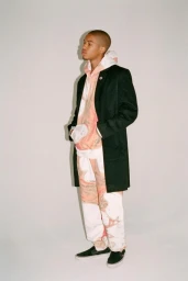 A young man wearing a black coat, patterned hoodie, matching sweatpants, and black slip-on shoes against a plain white backdrop.