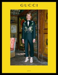Man wearing a dark green embroidered suit and bow tie standing in an ornate room with yellow border framing.