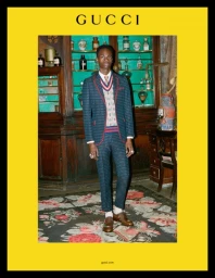 A young Black man wearing a patterned suit and sweater vest stands inside a vintage apothecary shop by Ari Marcopoulos.