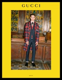 Young man in plaid coat and dark suit standing inside a historic room with wooden cabinets, framed by a bright yellow border with Gucci text