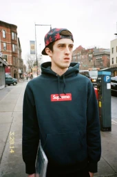 Young man wearing a dark navy hoodie with a red box logo and a printed cap standing on a New York city street