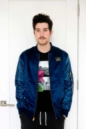 Man with curly dark hair and mustache wearing a blue satin bomber jacket standing indoors against a white door
