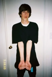 Young man in a black t-shirt standing indoors in New York by Ari Marcopoulos