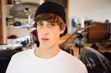 Young man wearing a white t-shirt and black cap indoors with drum kit in the background