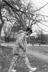 Monochrome photograph of a person walking through a park in camouflage jacket and pants beneath bare tree branches.