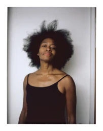 Portrait of a Black woman with voluminous afro hair wearing a black camisole against a plain white wall