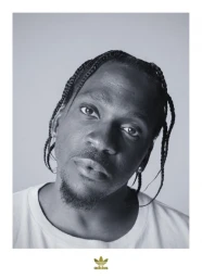Black and white portrait of a man with braided hair and a goatee wearing a white t-shirt against a light background.