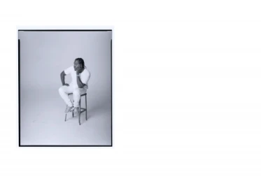 Portrait of a Black man seated on a stool wearing a white t-shirt and light-colored jeans against a white backdrop.
