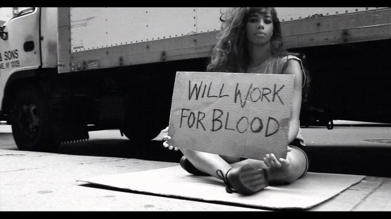 Monochrome image of a young woman sitting on cardboard holding a sign reading will work for blood in front of a truck