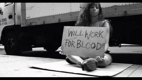 Monochrome image of a young woman sitting on cardboard holding a sign reading will work for blood in front of a truck