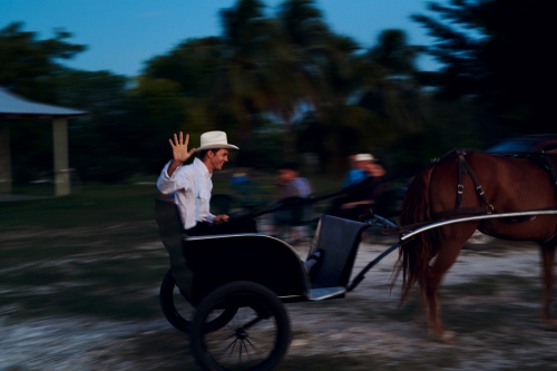 Man rides a horse-drawn buggy in motion with blurred background in an outdoor environment during dusk, wearing a white hat and light shirt.