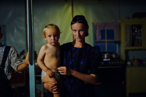 Young mother holding her child, both looking at the camera indoors. The photo evokes a sense of intimacy and quiet familial connection.