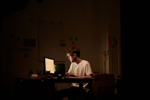 Man with glasses typing on a computer keyboard in a dimly lit room. Whiteboard with colorful sticky notes in the background.