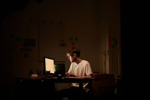 Man with glasses typing on a computer keyboard in a dimly lit room. Whiteboard with colorful sticky notes in the background.
